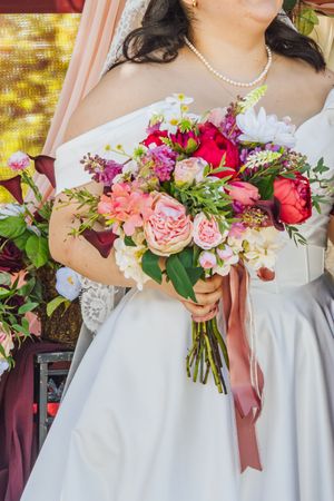 Bride in white gown holding a vibrant bouquet of pink, red, and white flowers with a mauve ribbon.