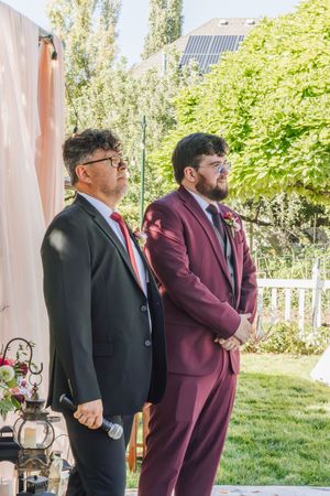Two men in suits stand at an outdoor wedding ceremony, one in black with red tie, one in burgundy with a boutonniere.