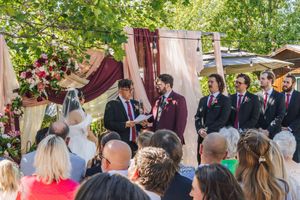 Outdoor wedding ceremony under a floral chuppah with groom in burgundy suit and groomsmen in dark attire.