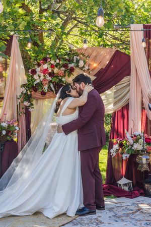 Bride and groom share first kiss at outdoor wedding altar adorned with burgundy drapes and floral arrangements.