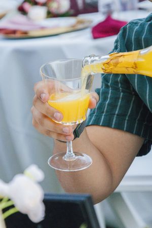Person pouring yellow juice from bottle into crystal wine glass at elegant outdoor event.