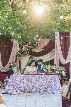 Bride and groom seated at floral sweetheart table under string lights and burgundy draping at outdoor wedding reception.