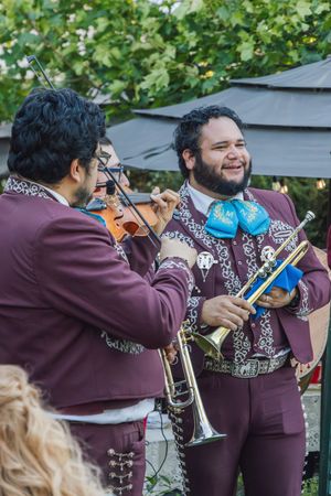 Two mariachi musicians in burgundy suits perform outdoors, one playing violin and one holding a trumpet.