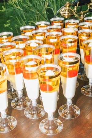 Elegant gold-rimmed champagne glasses filled with layered dessert featuring cream, orange jelly, and red fruit at an outdoor event.