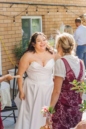 Bride in white strapless gown laughing with guest at outdoor wedding reception near brick building.