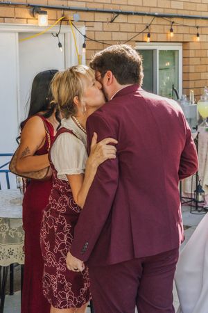 Couple sharing a kiss at an outdoor party, wearing matching burgundy outfits, with string lights and brick wall backdrop.