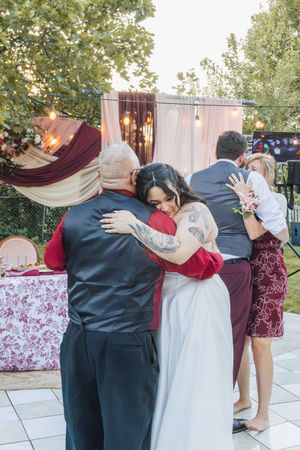 Bride in white dress hugs family members during outdoor wedding reception with string lights and floral backdrop.