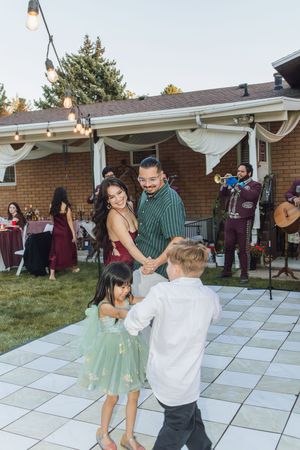 Family dancing together at an outdoor backyard wedding reception with mariachi band performing in background.