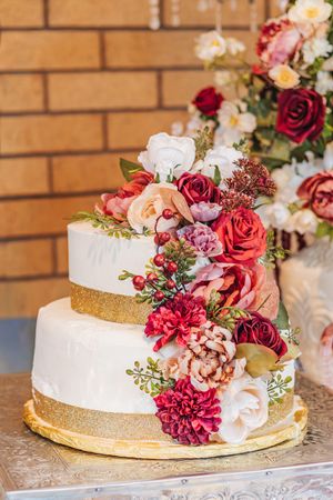 Three-tier white wedding cake with gold accents adorned with red roses, pink peonies, and white flowers.