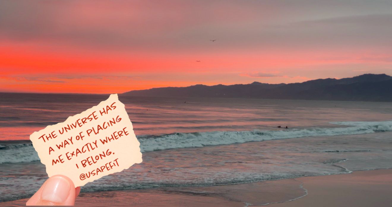 A hand holds a note reading a universe quote against a stunning pink and red sunset over a calm ocean beach.