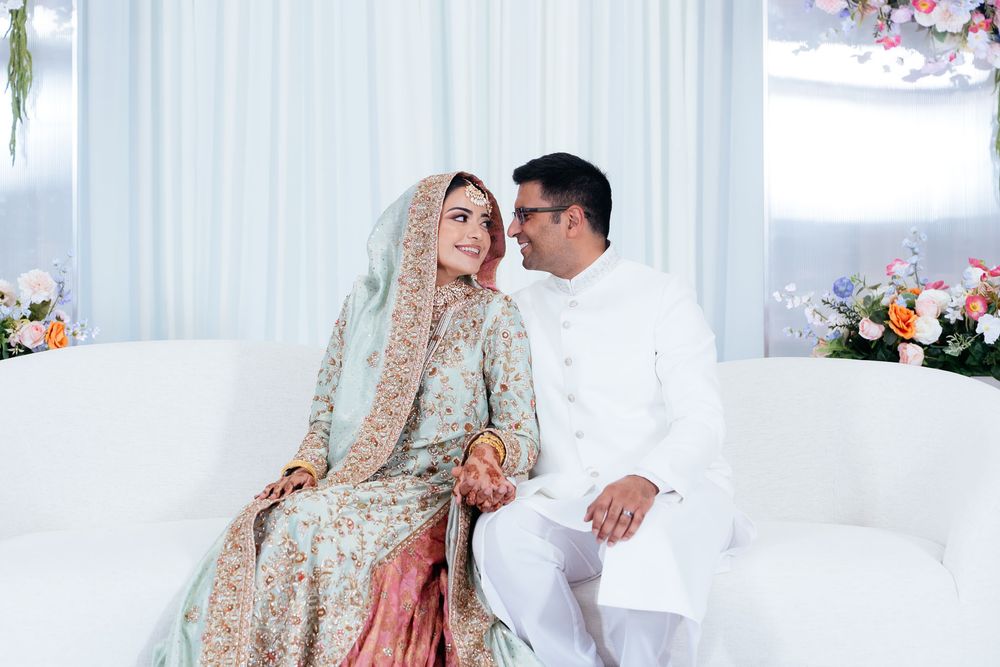 Smiling bride in an embroidered pastel wedding dress with dupatta sits beside her groom in a white sherwani on a white sofa among flowers.