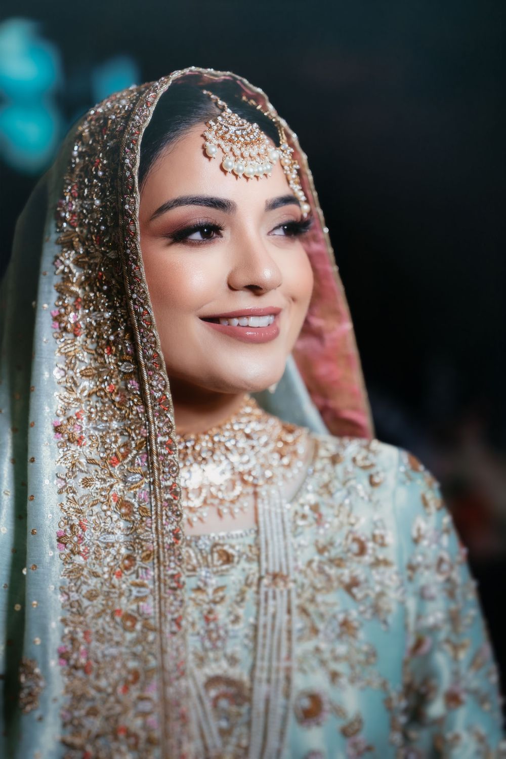 Smiling bride in a pastel blue embroidered dupatta and outfit with gold maang tikka and elegant jewelry, posing at a wedding.