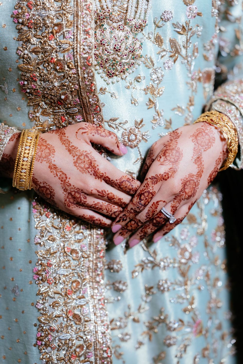 Bride's hands with intricate henna designs, gold bangles and a diamond ring, resting on a pale blue outfit with ornate gold embroidery.