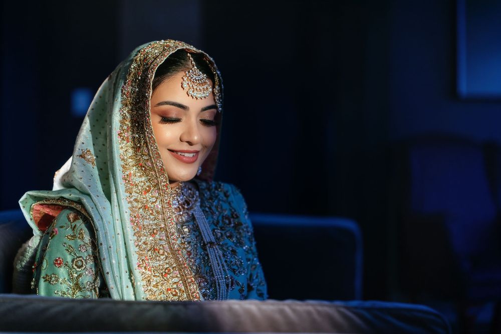 Smiling woman in an embroidered teal and gold dupatta with a forehead jewel, seated in soft blue light in traditional bridal attire.