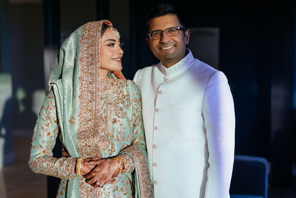 Smiling bride in an embroidered mint green and gold South Asian wedding dress with dupatta poses beside a groom in a white sherwani.