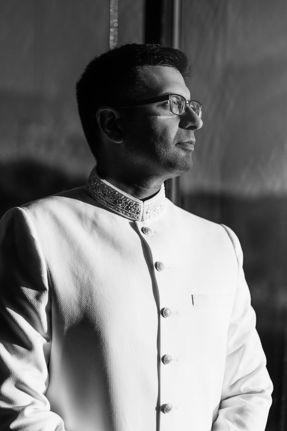 Black and white portrait of a man in glasses wearing a white embellished sherwani, gazing thoughtfully toward a window in soft light.