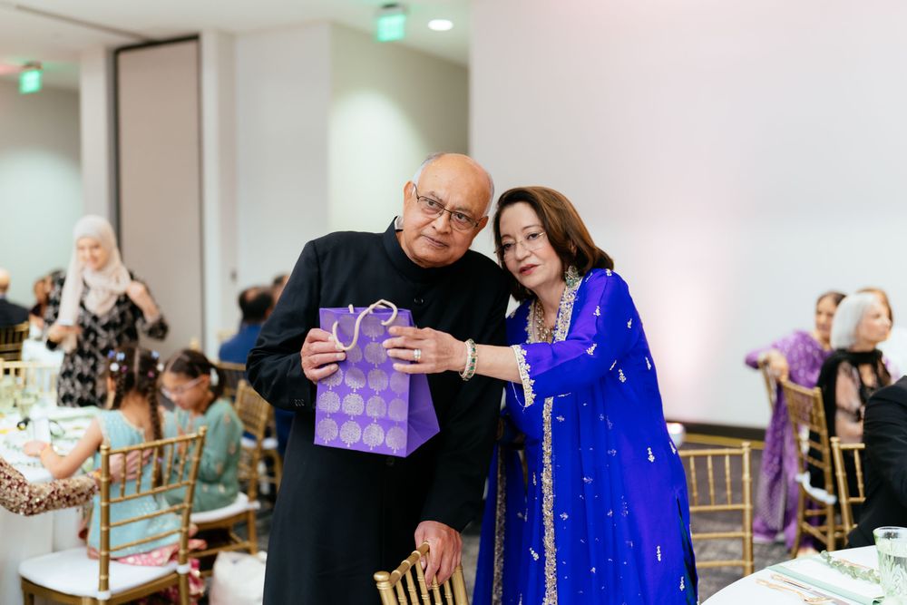 Smiling older man in a black outfit and woman in a royal blue embroidered dress hold a purple gift bag at a wedding reception.