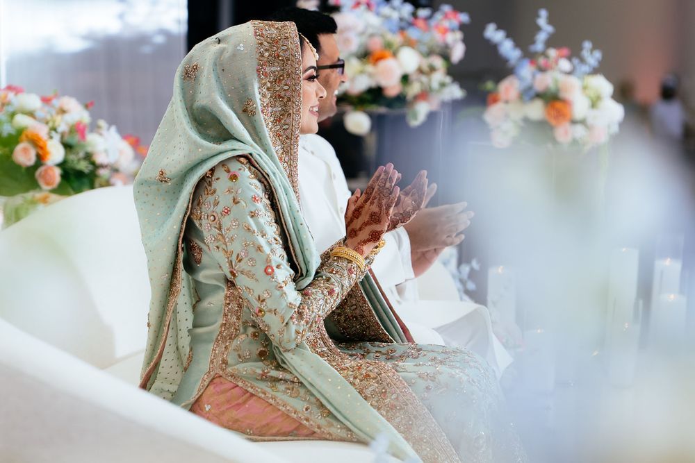 Bride in an embroidered sage green and gold outfit with a dupatta sits beside the groom, clapping at a floral-decorated wedding.