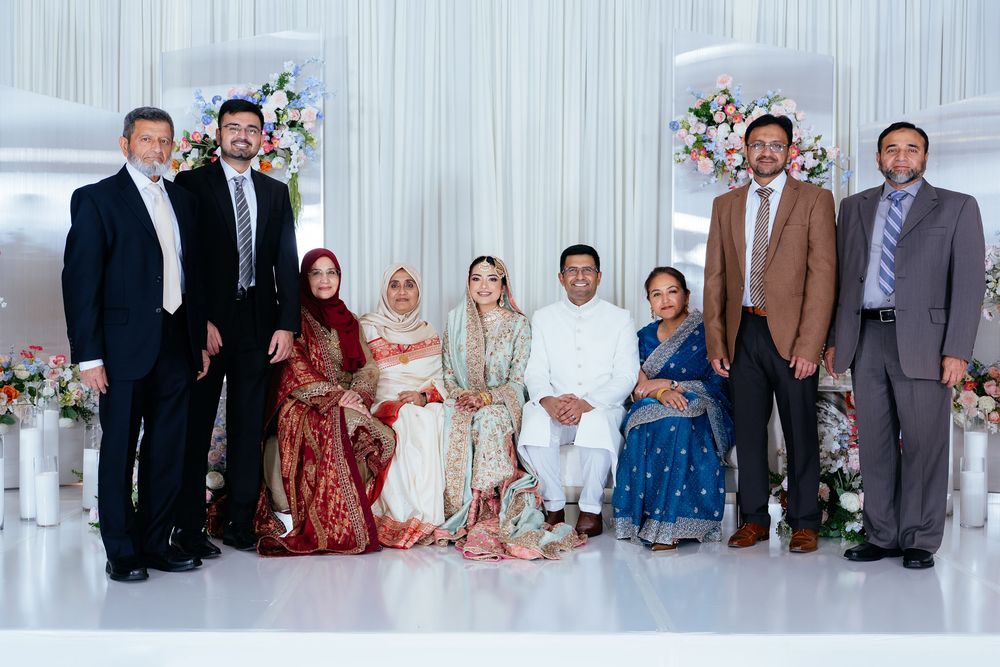Wedding party of nine in formal and traditional attire poses together on a white stage decorated with fresh flowers.
