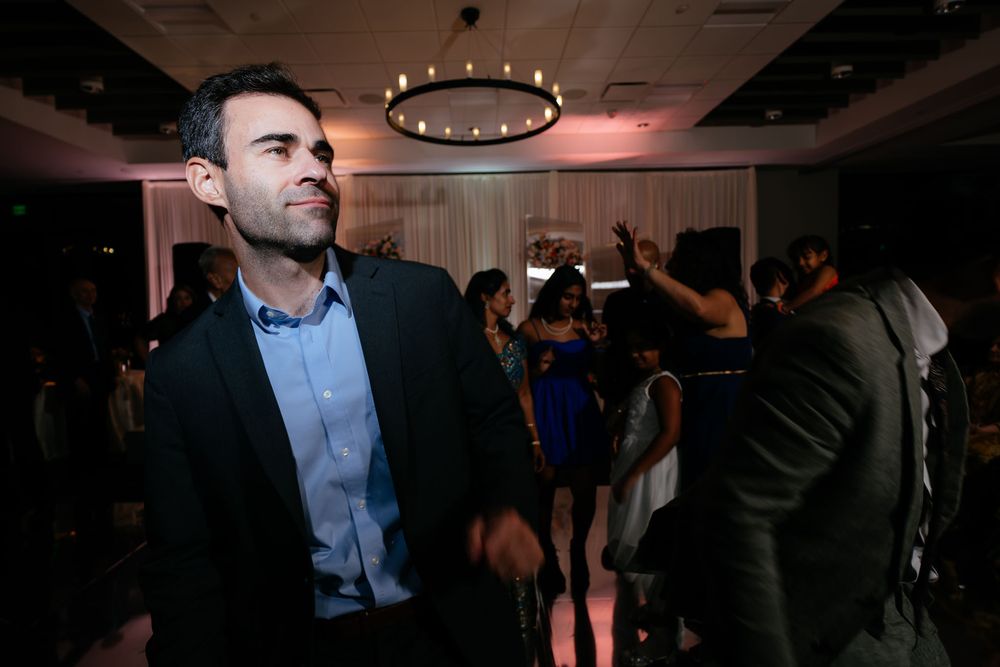 Man in a dark blazer looks off to the side while guests dance in the background at a lively evening wedding reception.