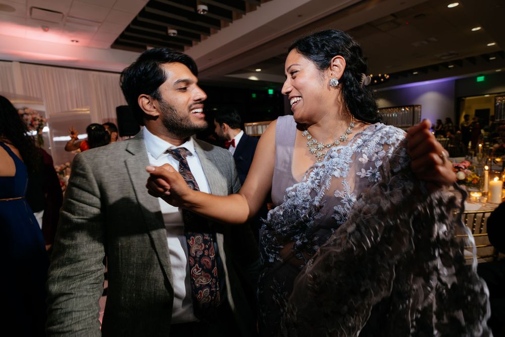 Laughing man in a gray blazer and patterned tie dances with a joyful woman in a sparkling gown at a lively wedding reception.