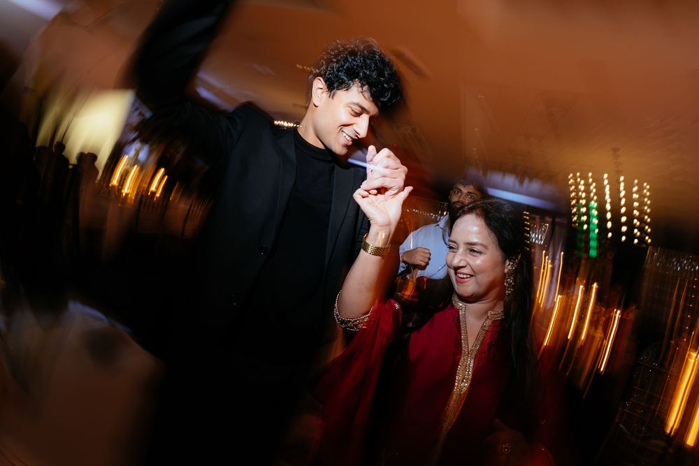 A smiling man in a dark jacket dances energetically beside a woman in red at a festive celebration with blurred, glowing lights.