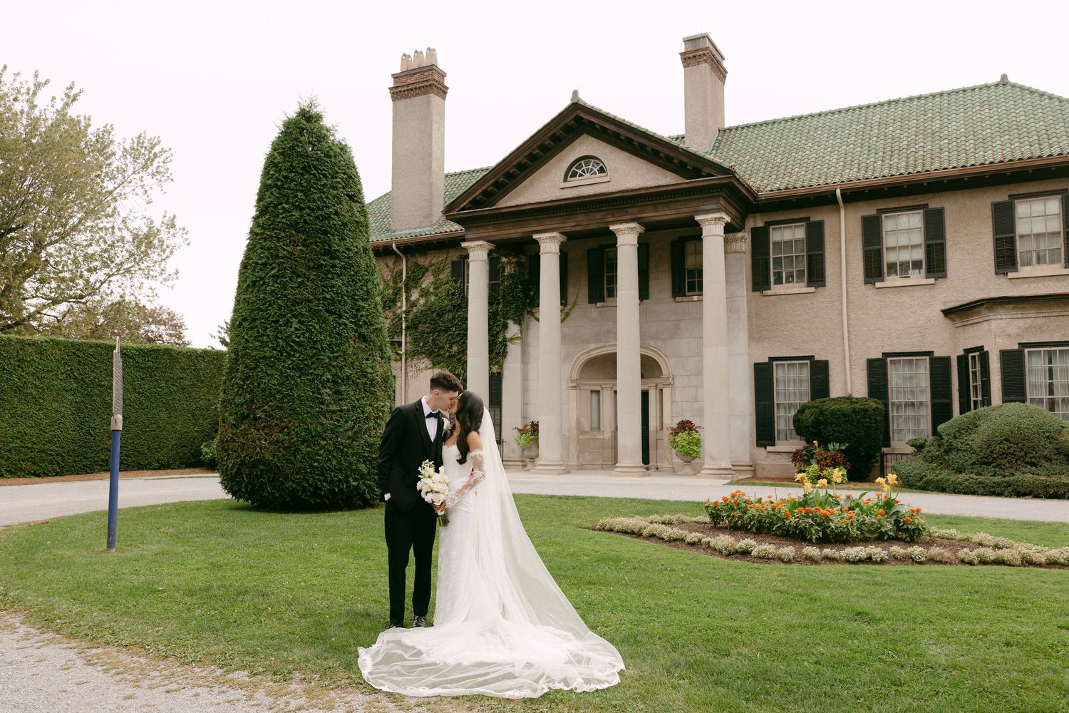 Bride and groom share a kiss on a manicured lawn in front of an elegant historic mansion with white columns on their wedding day.