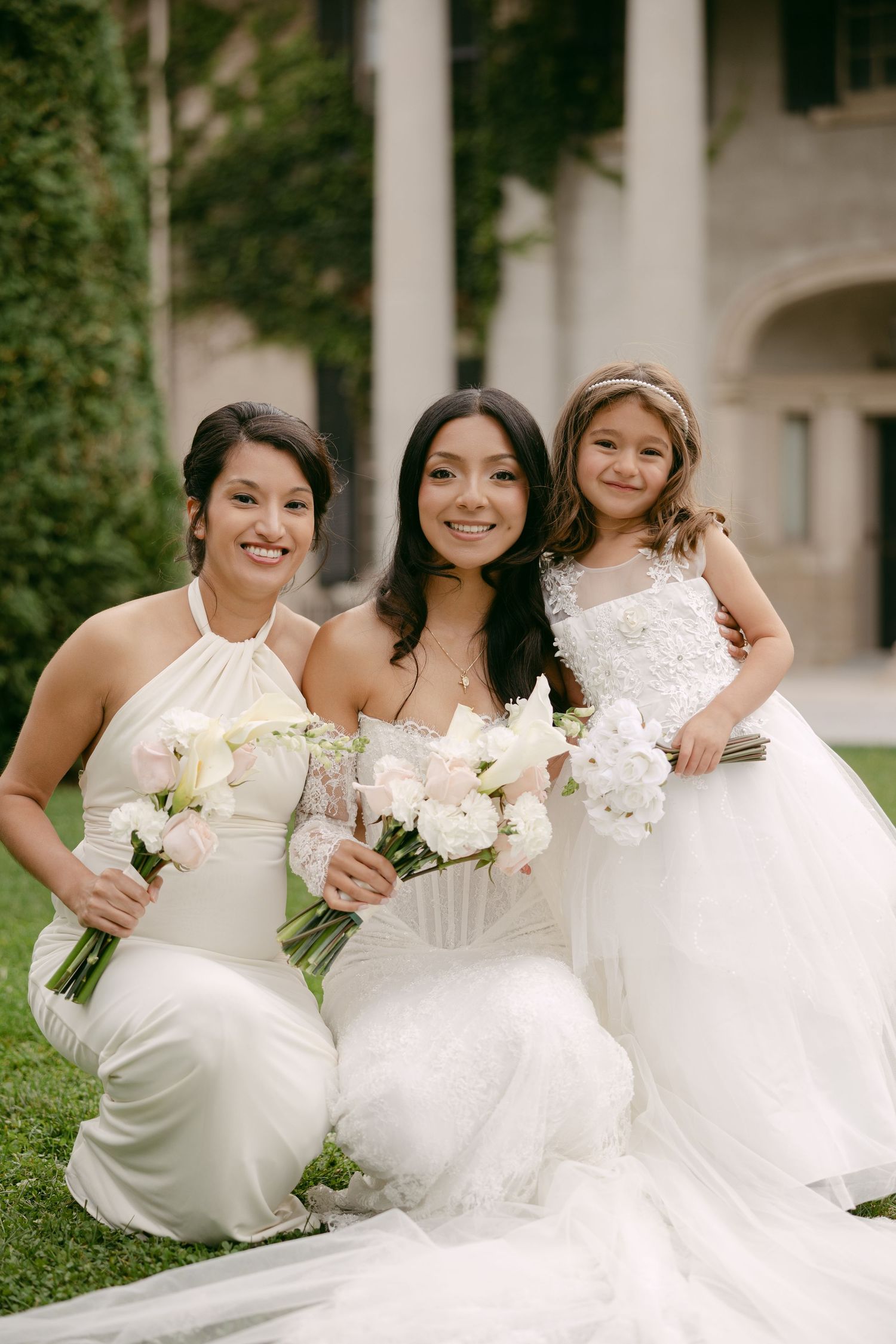 Smiling bride poses with a bridesmaid and flower girl holding white floral bouquets on a lush green lawn.