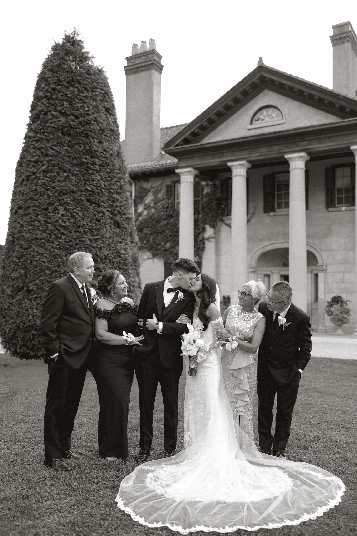 Newlyweds kiss in front of a grand columned mansion while both sets of parents smile beside them in a black and white photo.