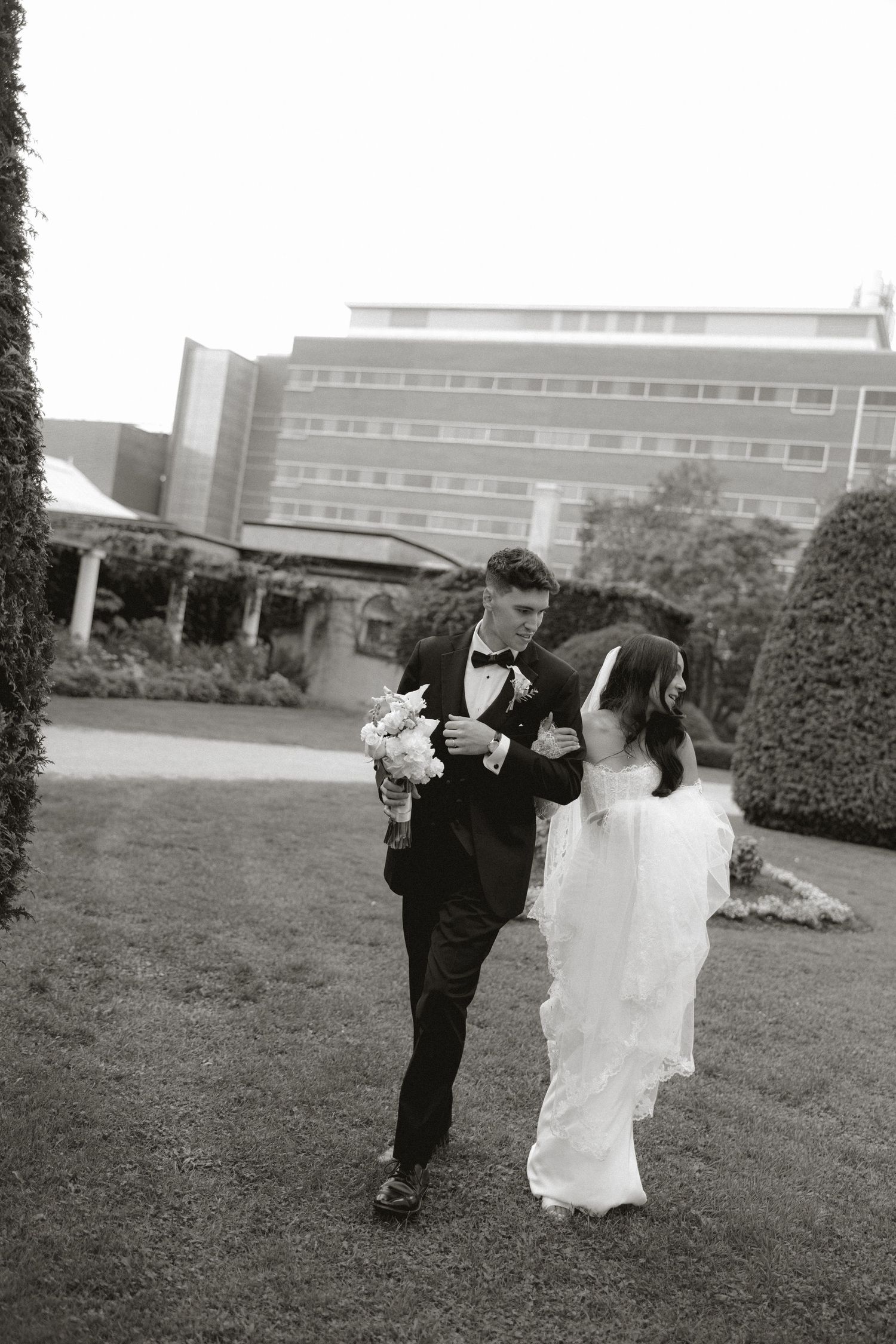 A bride and groom walk arm in arm across a lawn, the bride holding a white bouquet, in a black and white wedding photo.