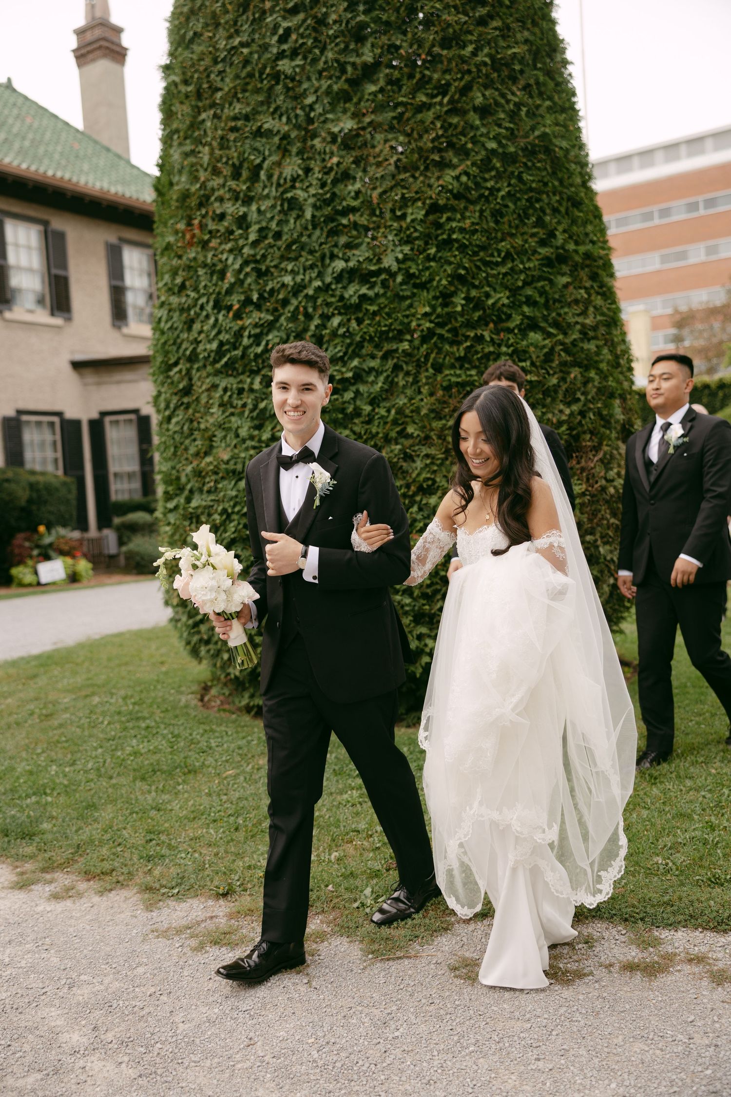 Smiling bride and groom with a white bouquet stroll past an ivy-covered hedge as groomsmen in black suits follow behind.