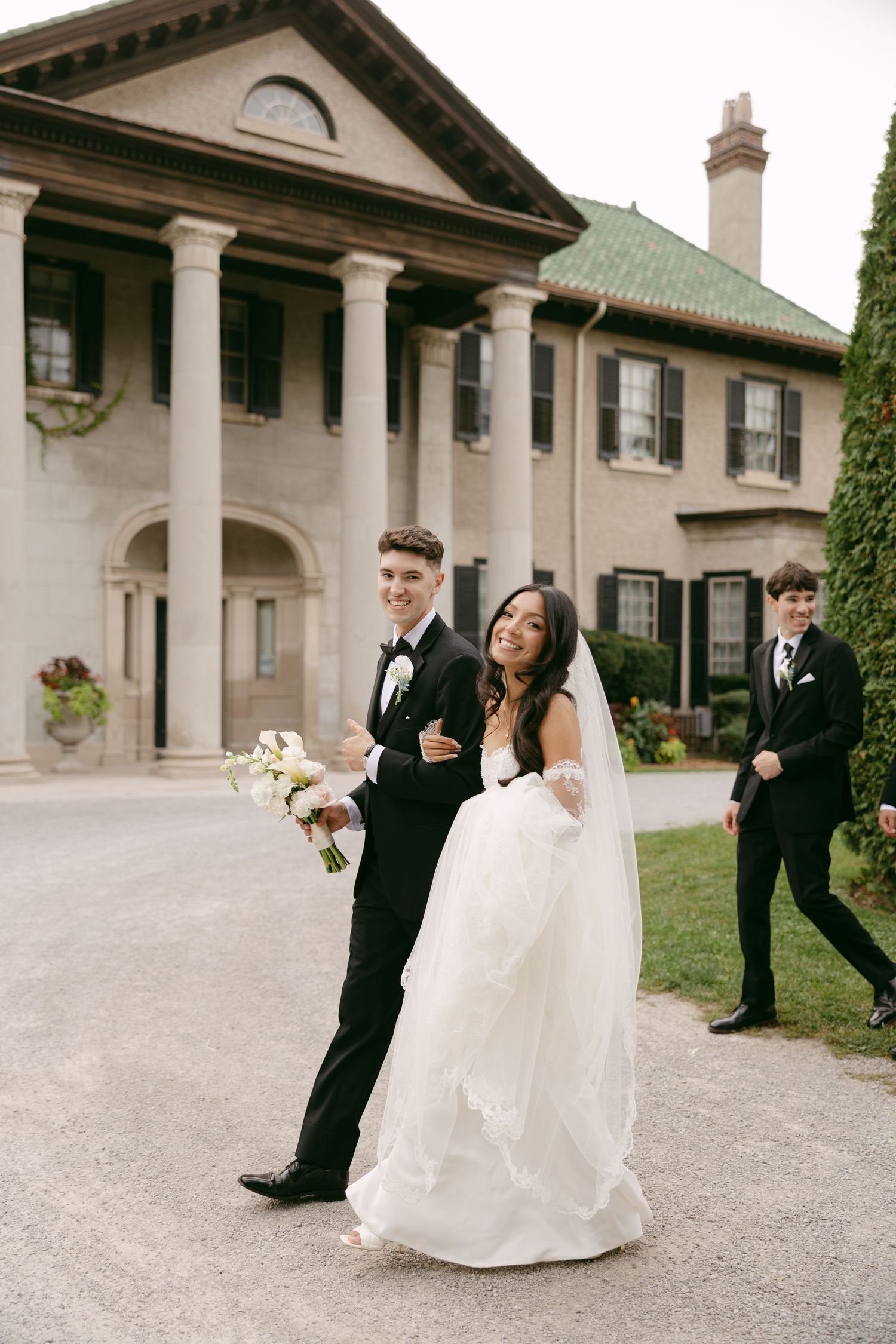 A smiling bride in a flowing white gown and veil carries a bouquet while walking with her groom outside a stately columned building.