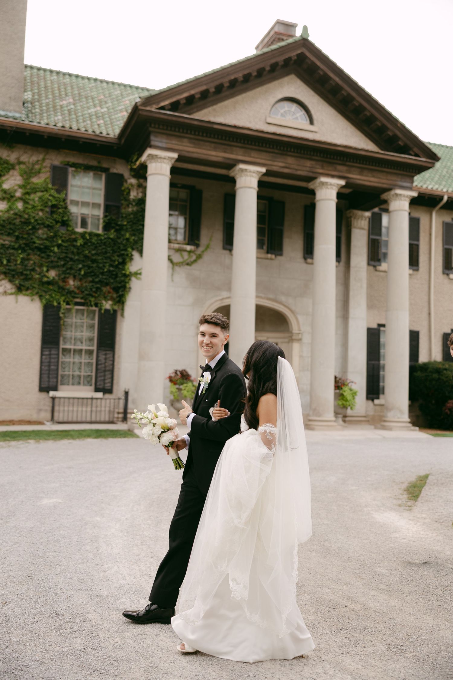 Bride in a white gown and veil walks arm in arm with a groom in a tuxedo holding a bouquet outside a grand columned estate.
