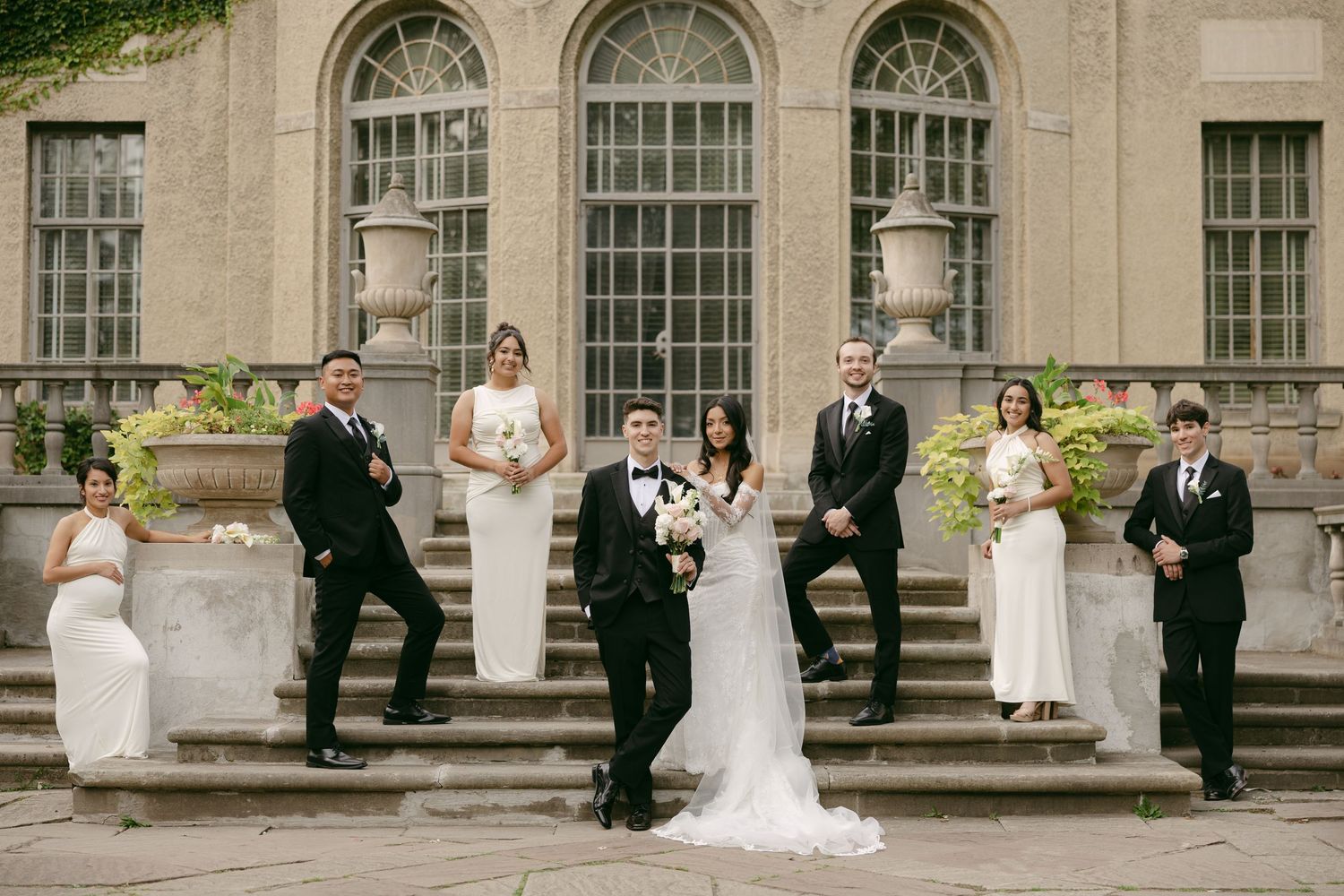 Wedding party in black suits and white dresses poses on stone steps outside a historic building with the bride and groom at center.