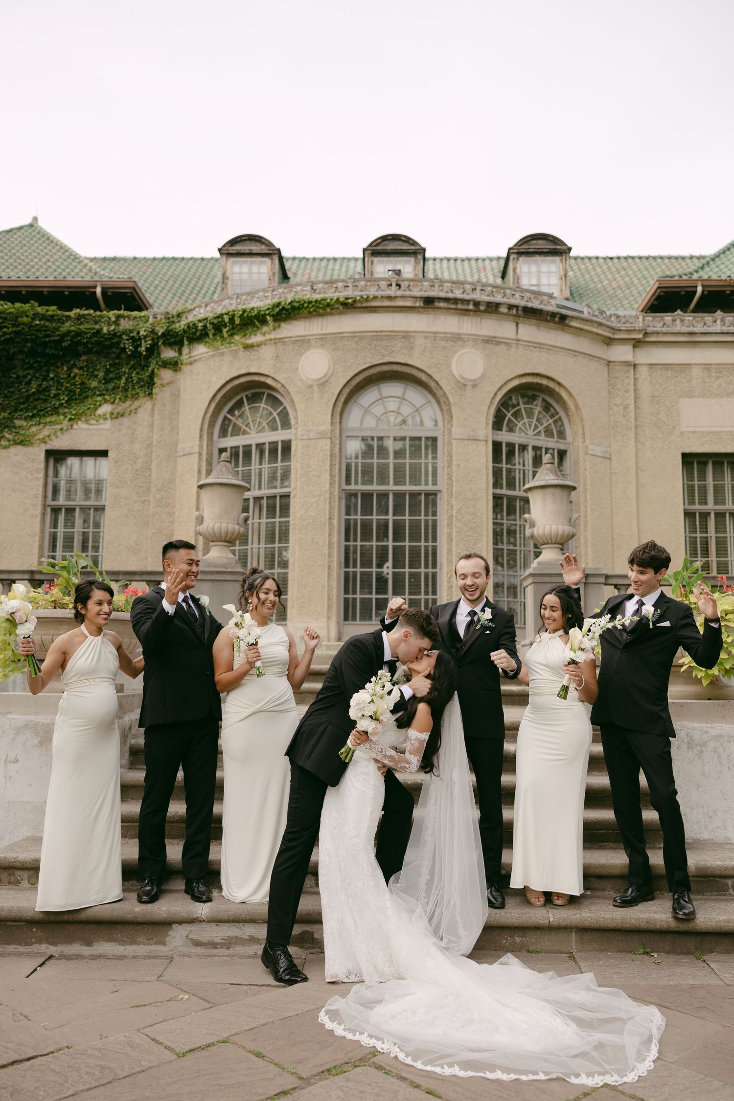 Bride and groom kiss on stone steps as the wedding party cheers in front of a historic arched estate.