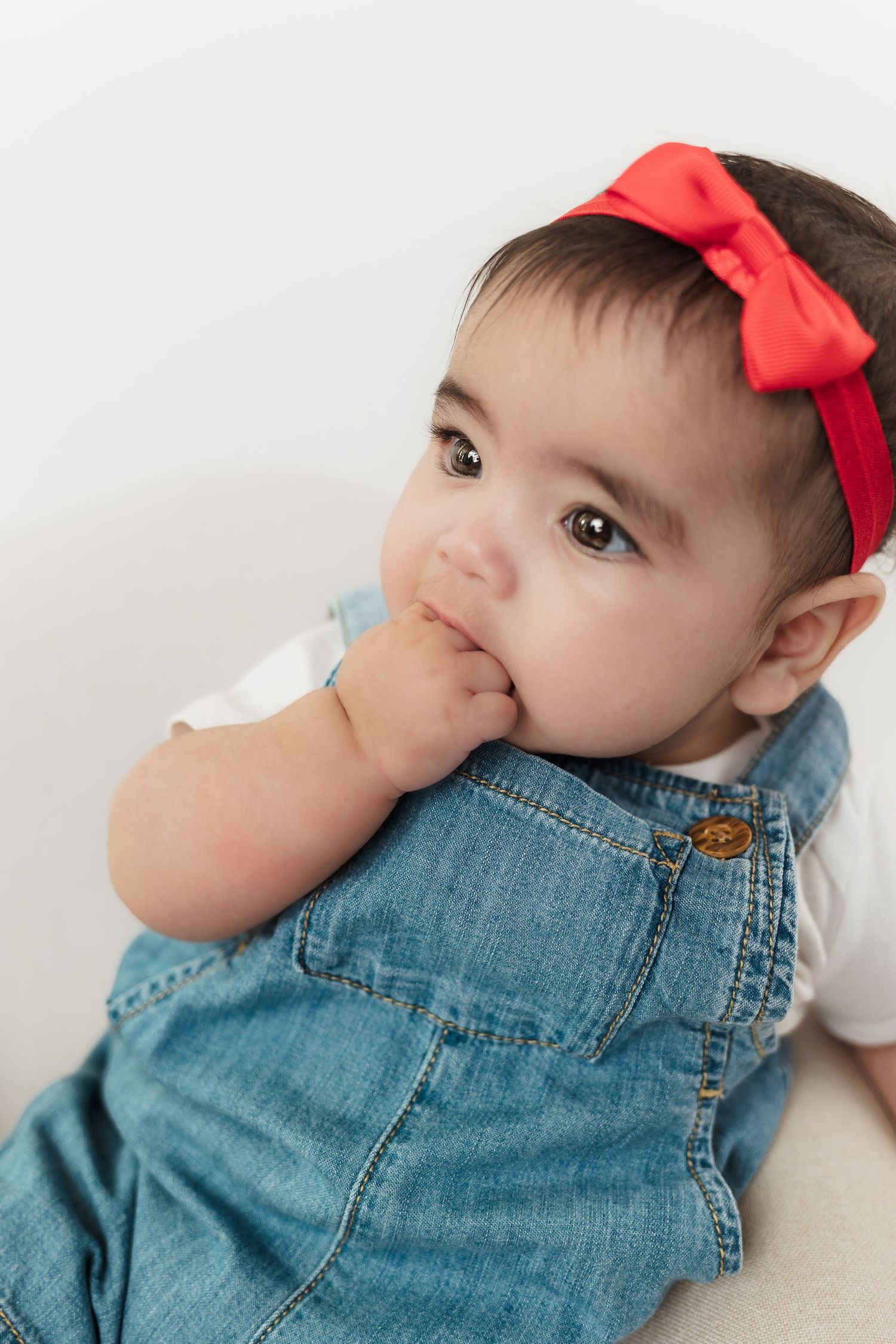 Adorable baby girl wearing denim overalls and a red bow headband, sitting and sucking her fingers against a white background.