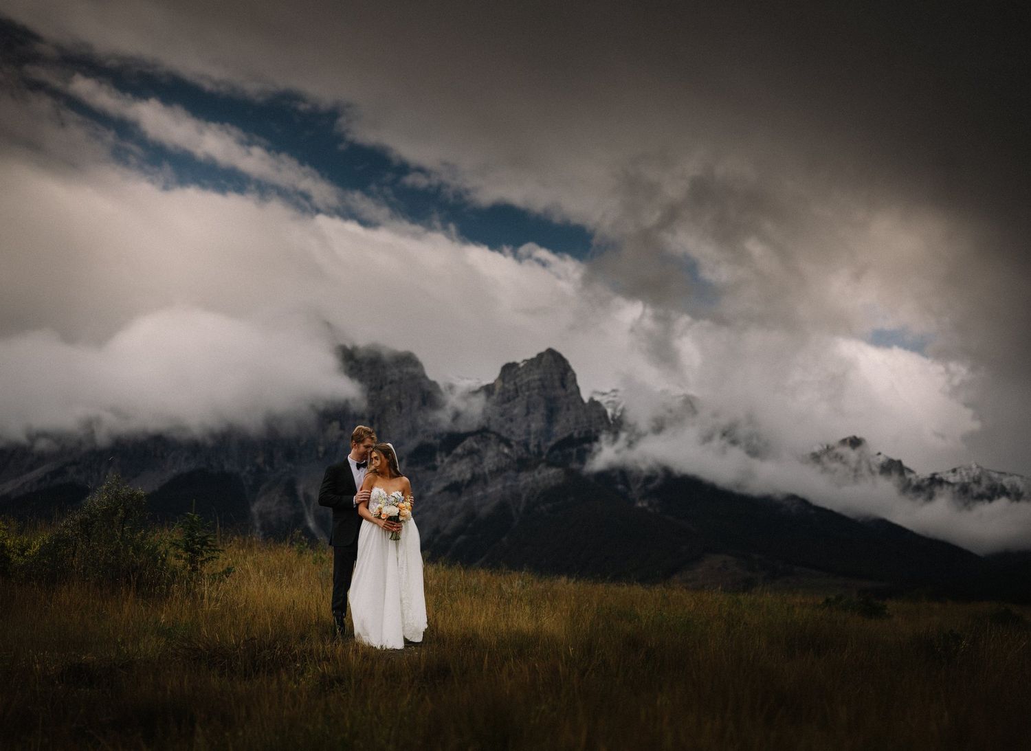 Bride and groom standing in a mountain field under dramatic storm clouds in the Canadian Rockies