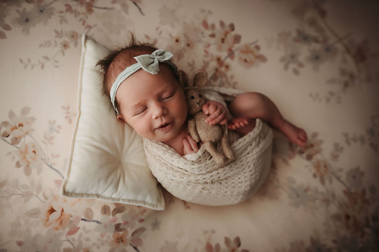 Sleeping newborn baby wrapped in cream knit swaddle with bow headband, holding stuffed animal on floral backdrop.