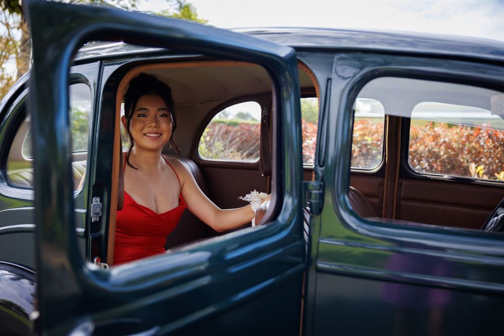 Smiling young woman in a red dress looks out the open window of a vintage dark green car, wearing a white corsage.