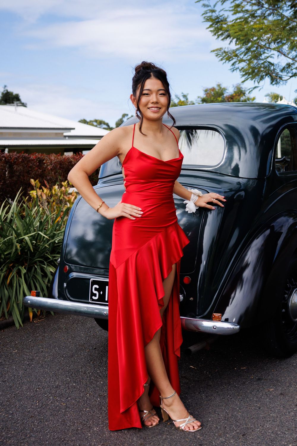 Young woman in a red satin high-low formal dress and strappy heels poses beside a vintage black car on a driveway.