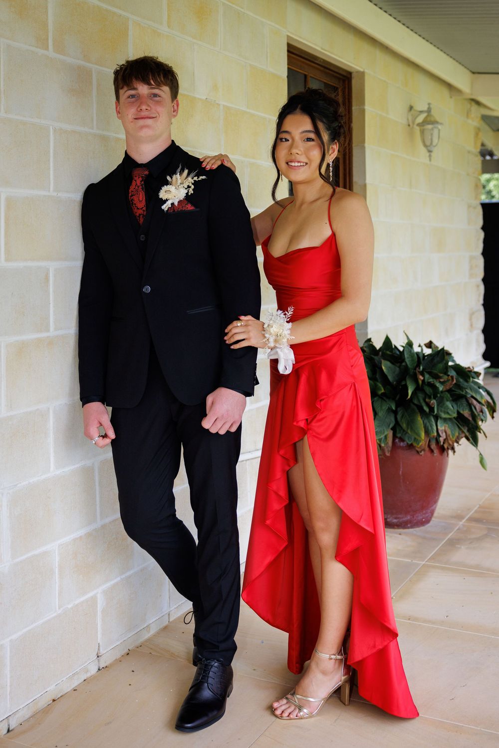 Young couple in formal wear pose against a sandstone wall, he in a black suit with a floral pin and she in a red slit gown.