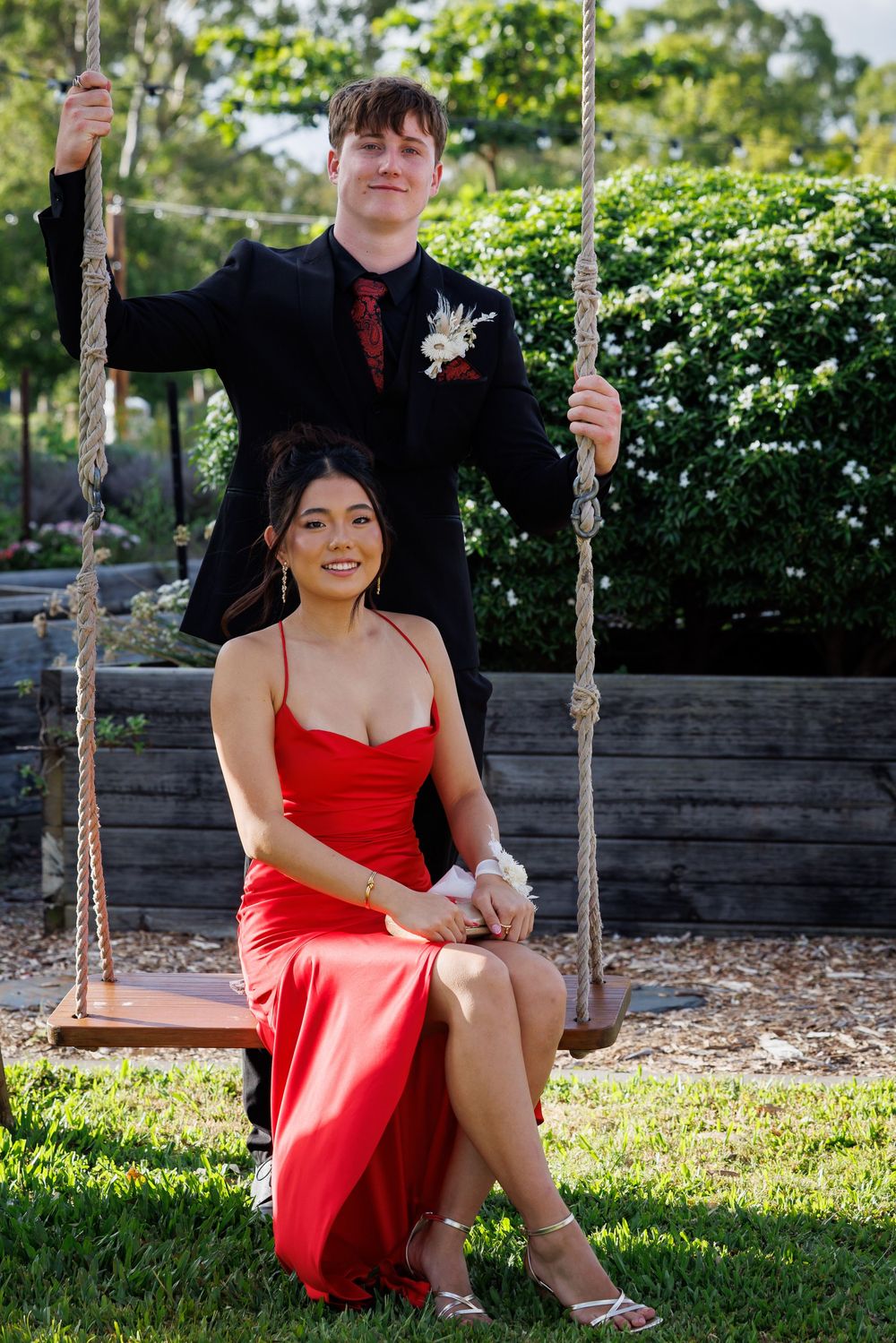 Young man in a black suit with a corsage stands behind a woman in a red satin dress seated on a rope swing in a garden.