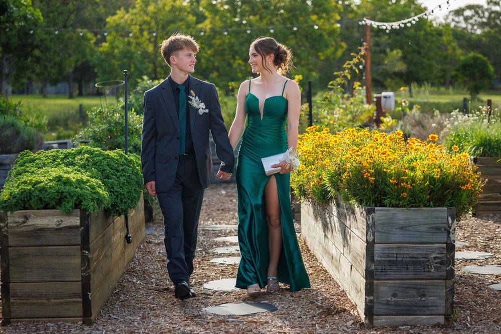Couple in formal attire, a young man in a dark suit and a woman in an emerald gown, walking hand in hand through a garden.