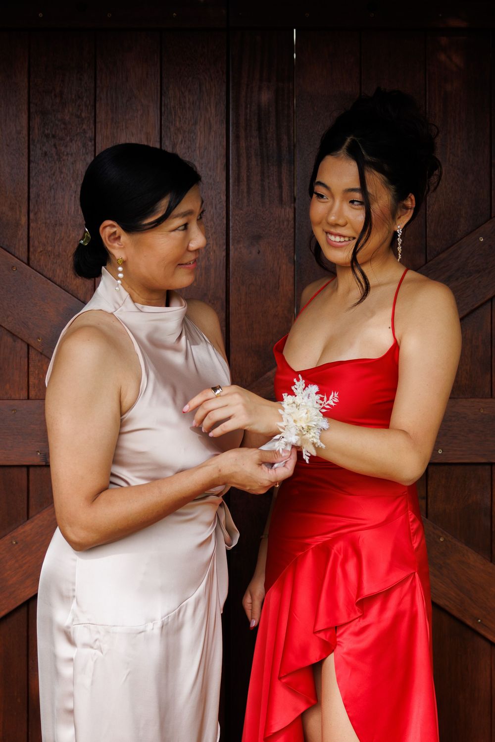 A woman in a satin champagne dress fastens a white floral corsage on the wrist of a young woman in a red dress by a wooden door.
