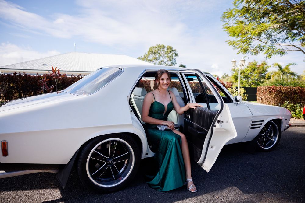 Young woman in an emerald green formal dress steps out of a classic white vintage car on a sunny day.