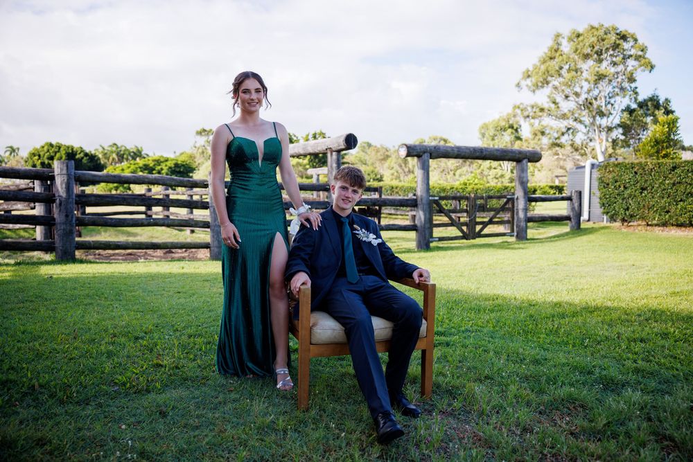 Young man in a suit sits on a wooden chair while a woman in a green gown stands beside him in front of a rustic timber fence.