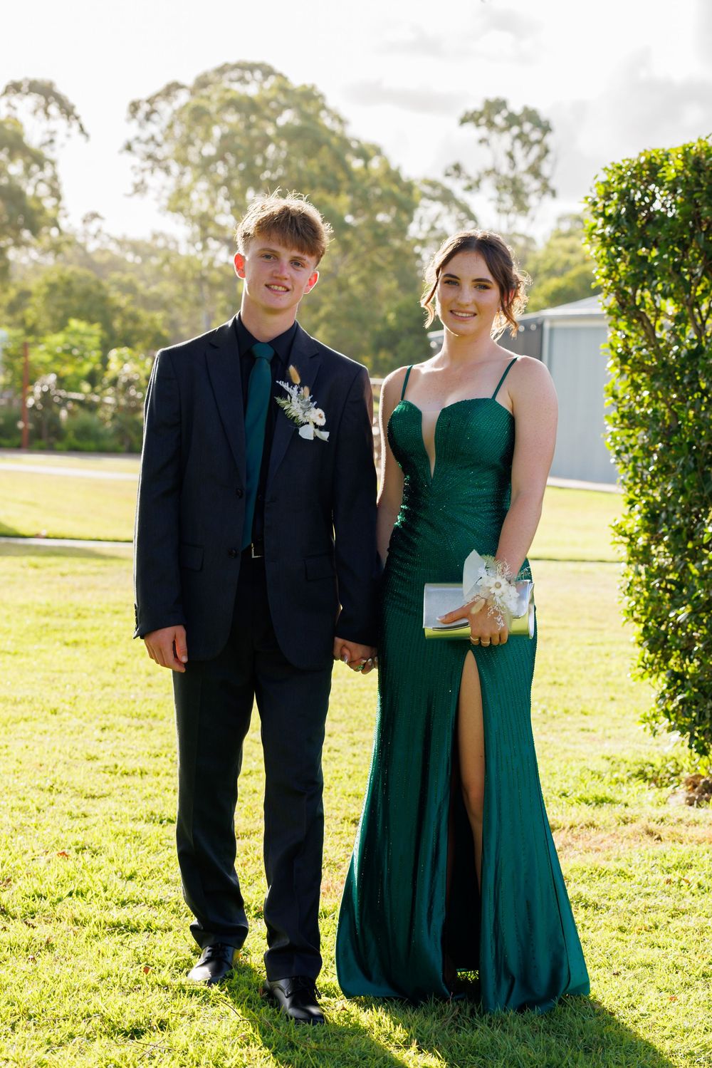Teen couple holding hands on a sunny lawn, the girl in a green gown with a high slit and the boy in a dark suit.