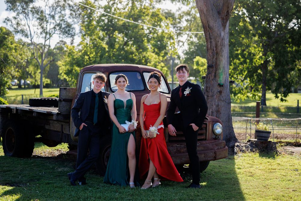 Two couples in formal wear, with dresses in green and red, pose beside a vintage truck under shady trees.