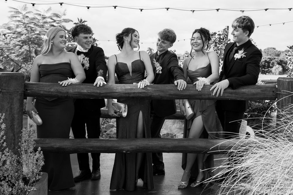 Black and white group photo of young friends in formal wear leaning on a wooden railing and laughing together outdoors.