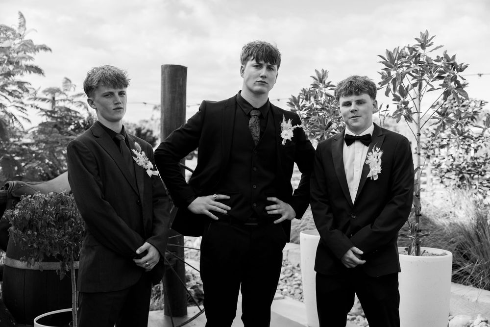Black and white photo of three young men in black formal suits and bow ties posing together near potted plants.